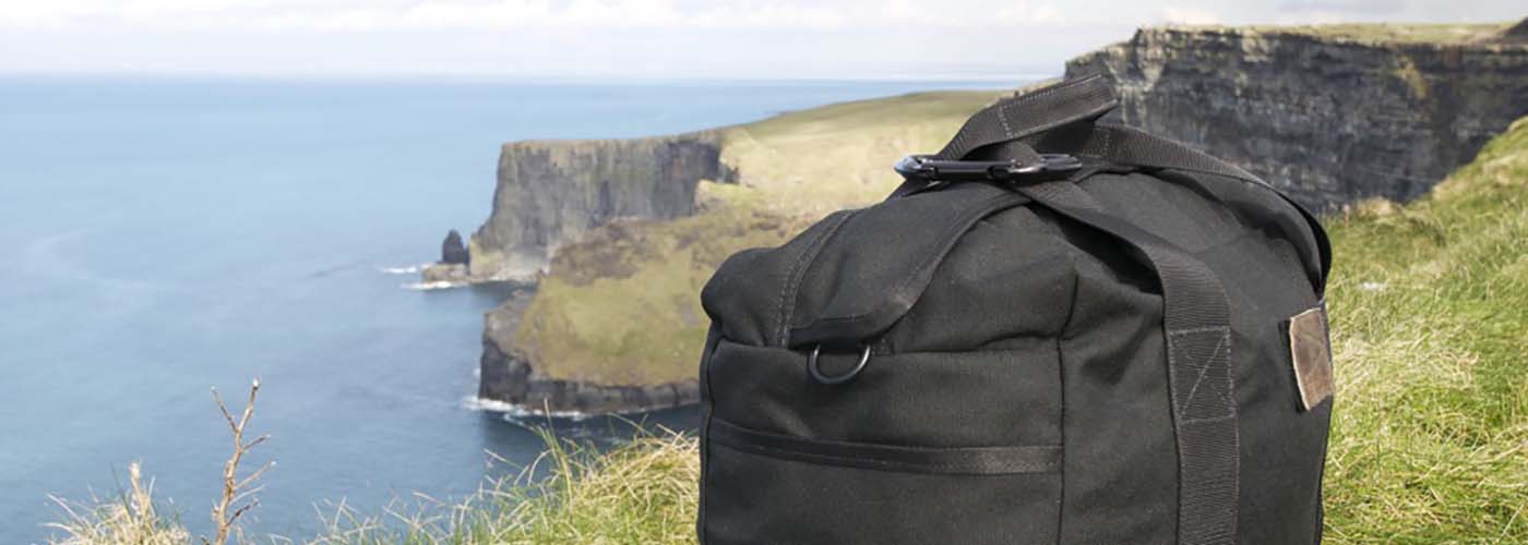 A black duffel bag on grass near the edge of a cliff overlooking the ocean, with towering cliffs in the background under a partly cloudy sky.