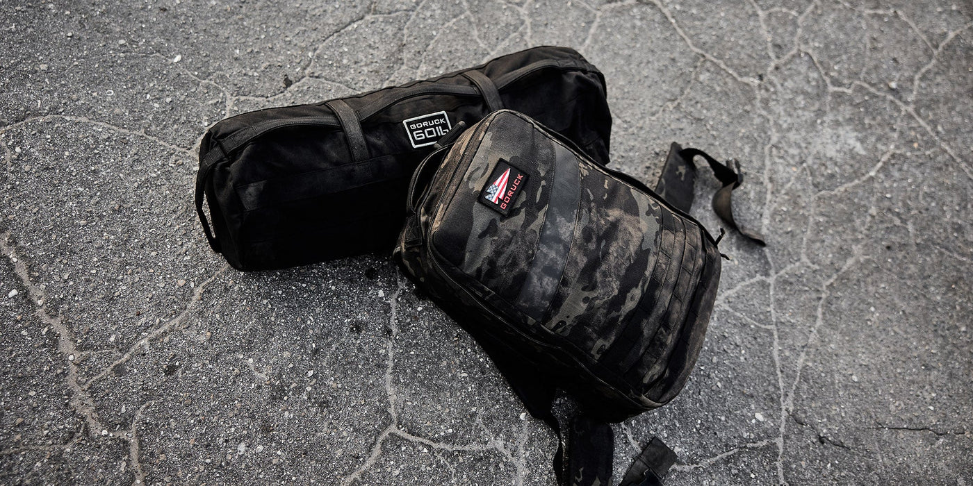 Two black duffle bags are placed on a cracked asphalt surface. One bag is labeled GORUCK 30L and the other has a camouflage pattern. Both bags are visibly worn and used.