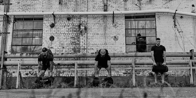 Three men lifting weights outdoors in front of an old brick building with pipework and large windows. The scene is black and white, with grass in the foreground.