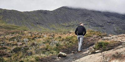 A person hikes up a rocky trail surrounded by a grassy landscape with mist-covered mountains in the background.