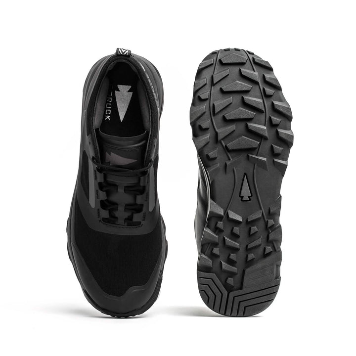 The image features a pair of Mackall shoes in Black + Forged Iron, from GORUCK. One shoe is upright, showcasing the top design, while the other lies on its side to emphasize the rugged sole with arrow-shaped tread patterns. These shoes are ideal for rucking or Special Forces training and seem designed for outdoor adventures.