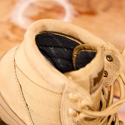 Close-up of the MACV-2 Safety Boot - Mid Top by GORUCK, showcasing its tan suede finish with laces, composite toe, and inner lining. The background reveals a blurred wooden surface adorned with subtle white circular patterns.