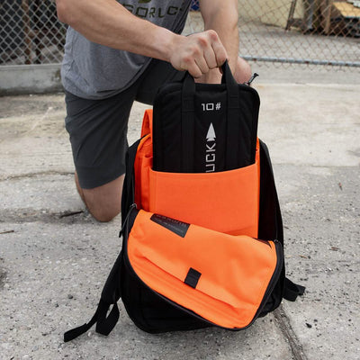 A person kneels on the ground, carefully placing a Sand Ruck Plate into an orange and black GORUCK backpack, which features a visible brand label. The scene unfolds outdoors near a chain-link fence, highlighting the rugged durability synonymous with a Lifetime Guarantee.