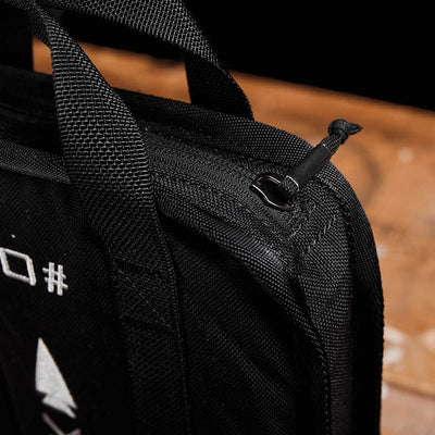 Close-up of a GORUCK Sand Ruck Plates fabric bag in black, featuring reinforced stitching and a visible zipper. The top handle and zipper pull can be seen, along with partial white text and symbols printed on the bag, guaranteeing quality to last a lifetime. The background is a wooden surface.