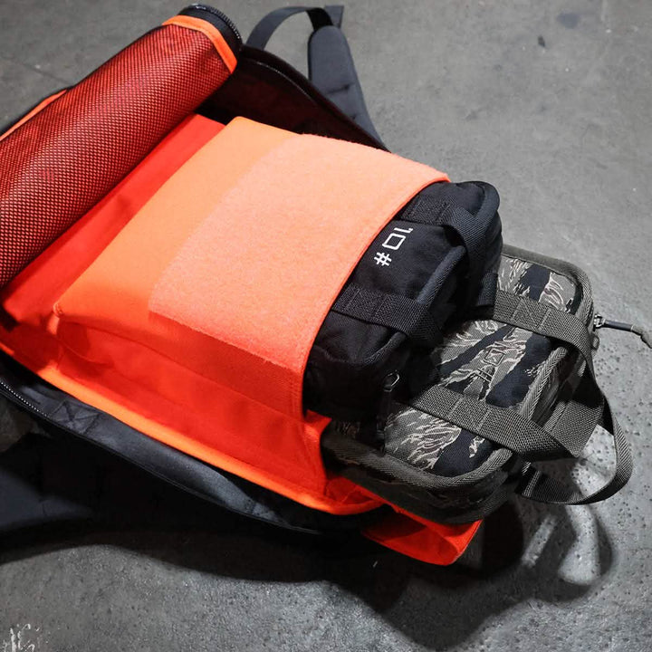 An open GORUCK Rucker backpack on a concrete floor displays its well-organized interior. It boasts a padded laptop compartment with an orange cover and includes a camo-patterned bag, perfect for carrying your essentials. This black backpack, enhanced with orange accents, offers a lifetime guarantee to ensure peace of mind.