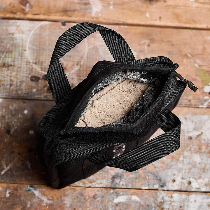 A black bag, reminiscent of a GORUCK with its sturdy handles and lifetime guarantee, lies open on a wooden surface. It reveals Sand Ruck Plates inside, slightly tilted to show the interior contents against the rustic wood background.