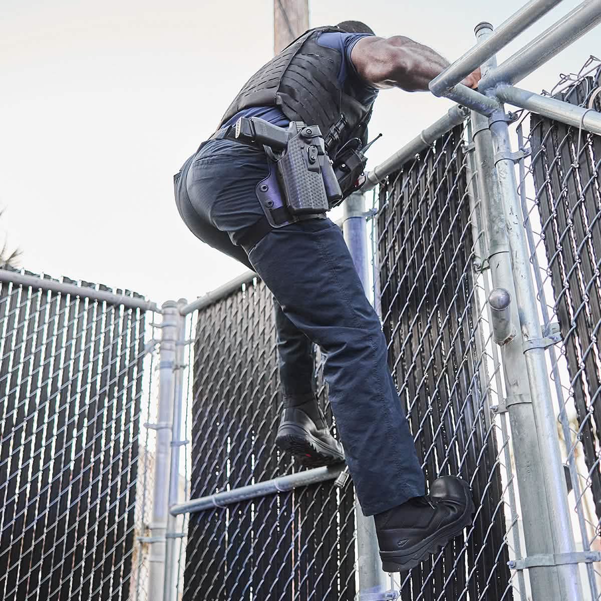 A person in a tactical uniform, conforming to Special Forces standards, climbs over a tall chain-link fence under the bright sun and clear sky. Dressed in dark clothing and wearing GORUCK's MACV-2 Safety Boot - High Top with a holstered gun, the scene exudes urgency and action.