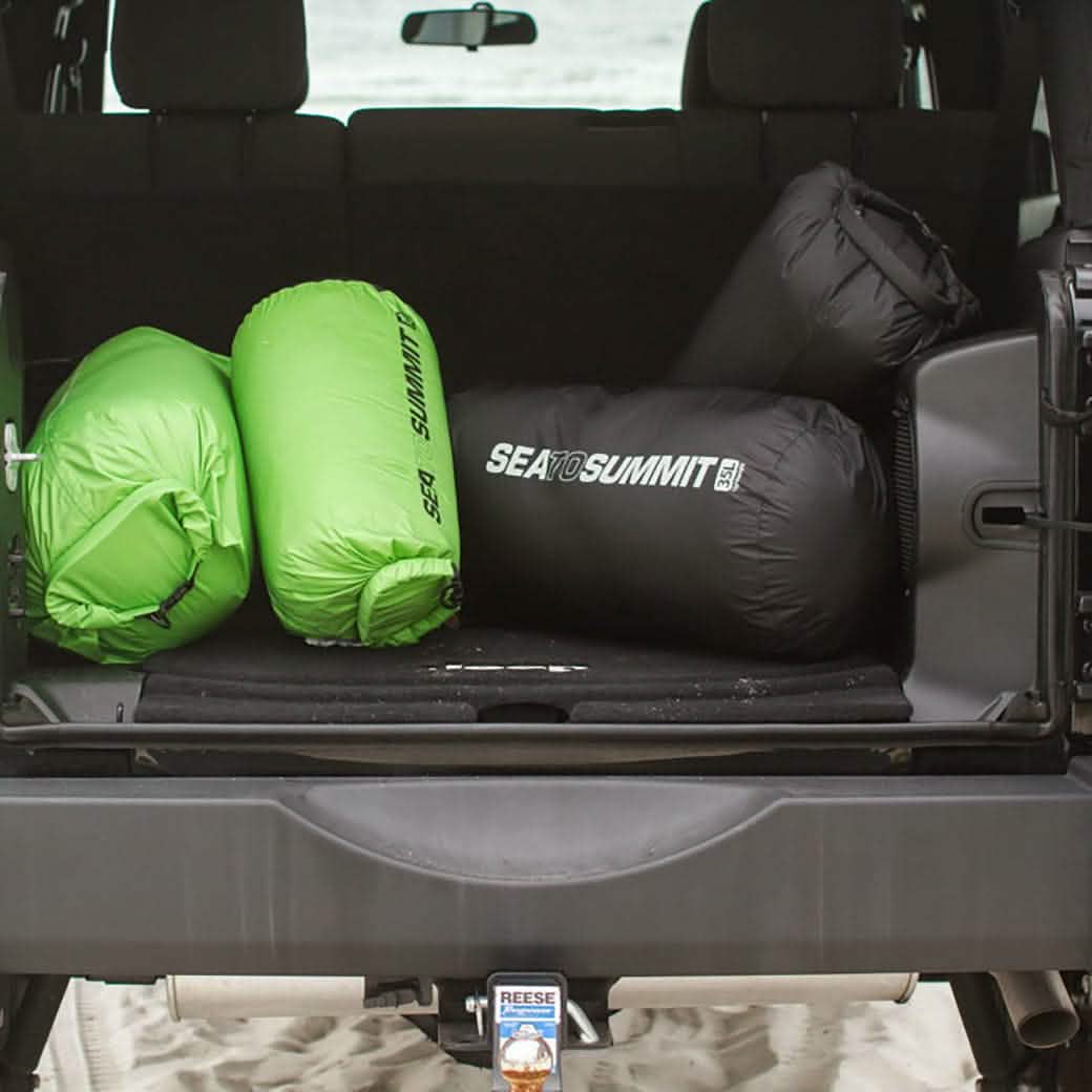 In the back of an open vehicle on a sandy surface, there are three Sea to Summit Dry Sacks. Two are green and one is black, all featuring a lightweight nylon design and waterproof closure. They are neatly arranged and ready for transport.