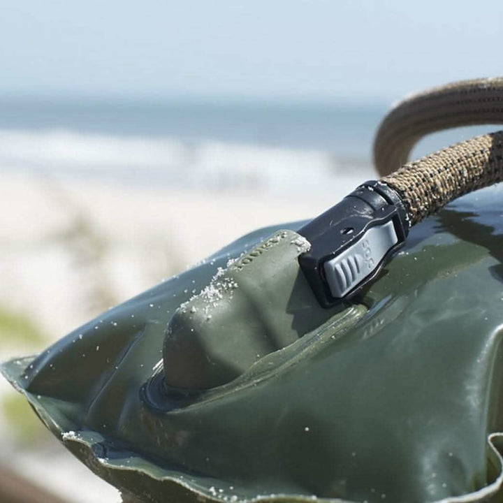 A close-up of the Source Hydration Bladder reveals its sand-covered green exterior, featuring a gray seal and a beige strap. Designed with antimicrobial technology, it is set against blurred ocean waves and a sandy beach backdrop, ideal for Special Forces who require efficient water solutions in coastal environments.