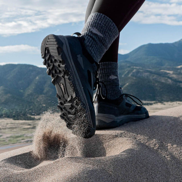 Close-up of a person wearing GORUCK's Mackall - Black + Forged Iron shoes paired with gray socks, stepping in sand on a dune. Sand is being kicked up by the shoes, with scenic mountains and a cloudy sky in the background.