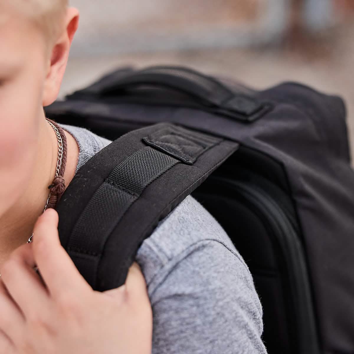 Close-up of a person in a gray shirt carrying the GORUCK KR1 kids rucksack with padded straps over their shoulder. The focus is on the KR1's strap and a bit of their face and hand, against a blurred background.