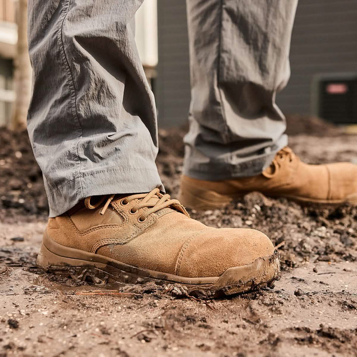 A person wearing the MACV-2 Safety Boot - Mid Top by GORUCK, featuring a composite toe and puncture-resistant sole, stands on muddy ground, hinting at outdoor or construction work. The focus is on the boots, which are slightly dirty, highlighting their rugged suitability for such settings.