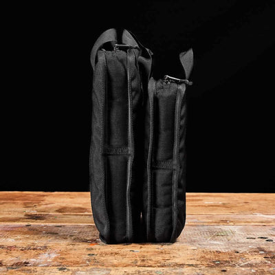 Two black fabric cases, one slightly taller and wider than the other, stand upright on a rustic wooden surface against a black background. Their sturdy design mirrors the resilience of Sand Ruck Plates by GORUCK, providing sleek functionality and the assurance of enduring quality.