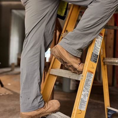 A person wearing MACV-2 Safety Boots - Mid Top by GORUCK and gray pants stands on a partially visible yellow ladder in a dimly lit room under construction, with wooden beams and tools in sight.
