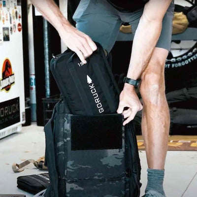 A person in a gray shirt and shorts is inserting a GORUCK Sand Ruck Plate, engineered for secure fitting, into a camo-patterned backpack. The indoor setting showcases various posters and gear, emphasizing the GORUCK brand's lifetime guarantee for the Rucker's durability and quality.