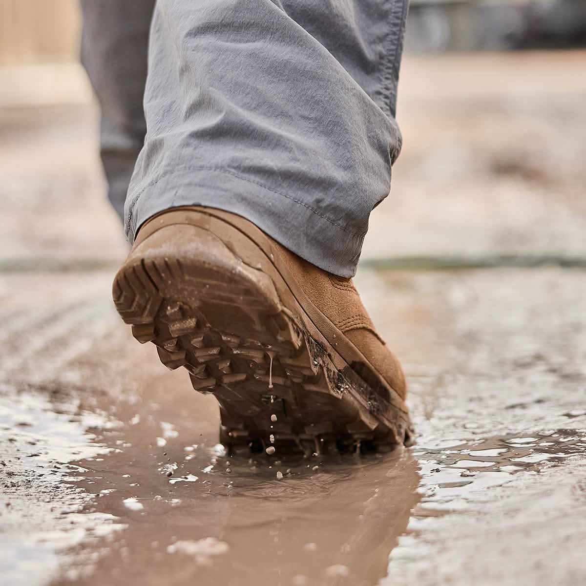 A close-up shows a person wearing GORUCK's MACV-2 Safety Boot - Mid Top in beige, featuring composite toes, as they walk through a shallow puddle on a muddy surface. The textured, puncture-resistant soles appear visibly wet, suggesting rugged outdoor activity. The person is dressed in gray pants.