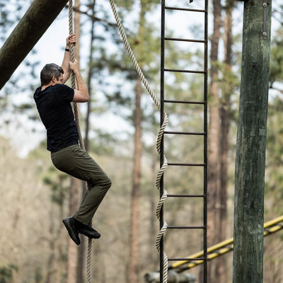 Wearing the GORUCK Mackall in Black + Forged Iron, a man scales a rope on an outdoor obstacle course that echoes Special Forces training. Beside him stand a wooden pole and metal ladder, set against a backdrop of trees.