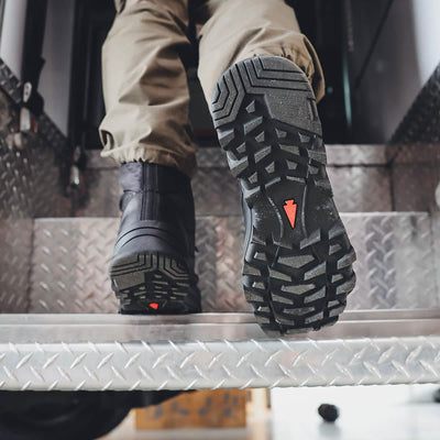 A close-up shows a person's feet resting on metal steps, clad in GORUCK's MACV-2 Safety Boot - High Top, featuring rugged black material with deep treads and red accents. Paired with tan pants, this ensemble exudes a utilitarian and industrial vibe, meeting Special Forces standards for durability and performance.