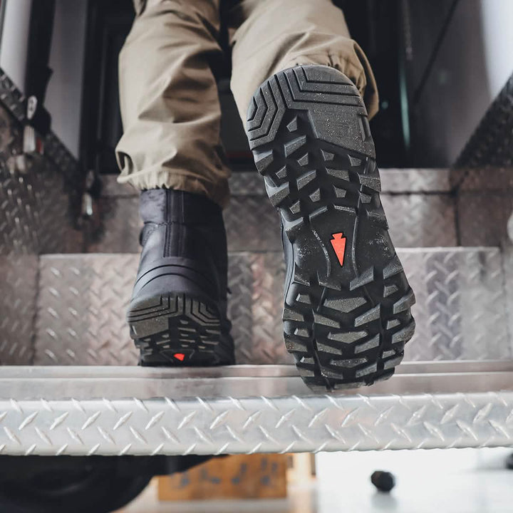 A close-up shows a person's feet resting on metal steps, clad in GORUCK's MACV-2 Safety Boot - High Top, featuring rugged black material with deep treads and red accents. Paired with tan pants, this ensemble exudes a utilitarian and industrial vibe, meeting Special Forces standards for durability and performance.
