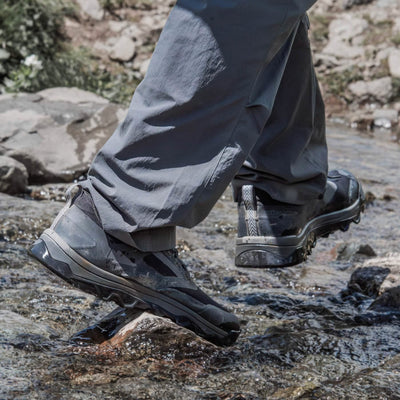 A person in gray pants crosses a small rocky stream, wearing GORUCK's Mackall shoes in Black + Forged Iron. The water is clear, revealing a few small rocks in this serene outdoor setting, reminiscent of Special Forces training grounds.