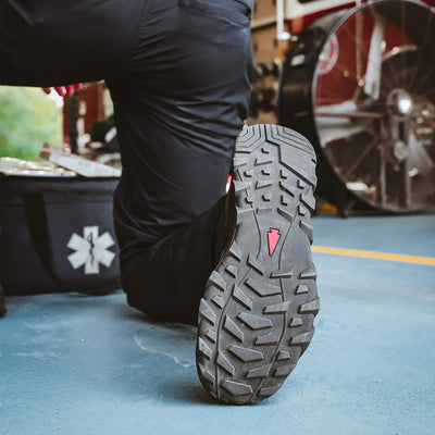 Close-up of a person kneeling on one knee, emphasizing the tread of the MACV-2 Safety Boot - High Top by GORUCK, featuring a red triangle logo. The background displays a fan, a window with greenery, and a black bag adorned with a medical symbol—crafted to meet Special Forces standards.