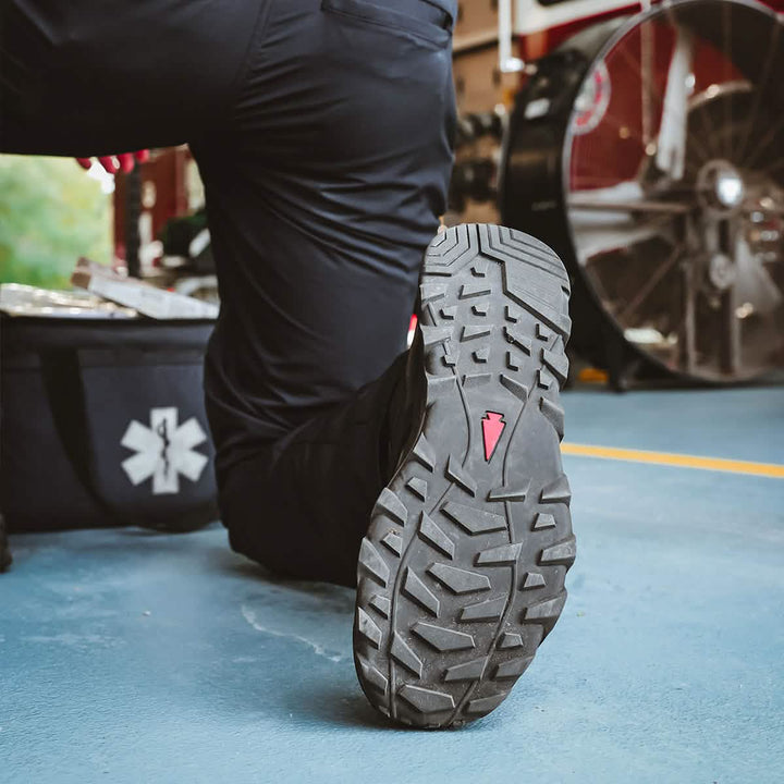 Close-up of a person kneeling on one knee, emphasizing the tread of the MACV-2 Safety Boot - High Top by GORUCK, featuring a red triangle logo. The background displays a fan, a window with greenery, and a black bag adorned with a medical symbol—crafted to meet Special Forces standards.