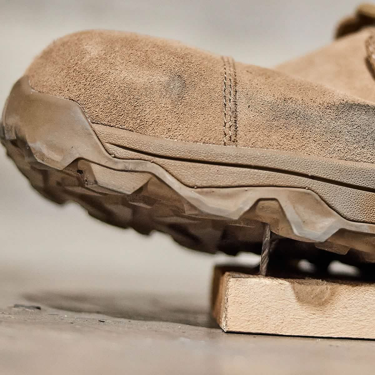 Close-up of the MACV-2 Safety Boot - Mid Top by GORUCK resting on a wooden block. The brown hiking boot, similar to rugged work boots, features a puncture-resistant sole marked with visible dirt and wear, emphasizing its outdoor functionality. The image highlights the suede material's texture and the tread pattern of the sole.