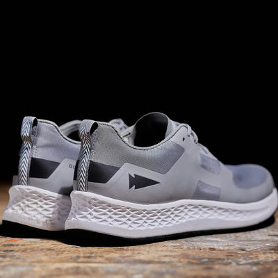 A pair of sleek Women's Rough Runner - Speed Grey + White shoes by GORUCK, featuring white EVA midsoles and black accents, sits on a wooden surface against a dark background. The heel tab boasts a zigzag pattern for enhanced style and gradient density, offering both comfort and performance.