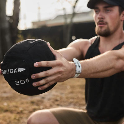 An individual sporting a cap and a fitness watch is outdoors, potentially at a park, intensely exercising with a black GORUCK Sand Medicine Ball to enhance their core stability.