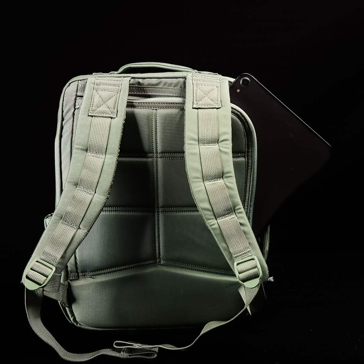 A GORUCK KR1 kids rucksack in green, featuring padded straps and visible stitching against a black background. A black tablet partially sticks out from a side pocket, making it perfect for carrying school supplies.