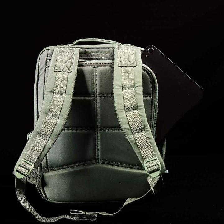 A GORUCK KR1 kids rucksack in green, featuring padded straps and visible stitching against a black background. A black tablet partially sticks out from a side pocket, making it perfect for carrying school supplies.