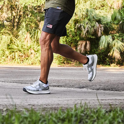 Jogging along a paved road enveloped by lush greenery, the individual sports high mileage running shoes featuring GORUCK's Men's Rough Runner design in White + Speed Grey. They are dressed in black shorts with an American flag patch and a green shirt, while trees and bushes create an ideal scenic backdrop.