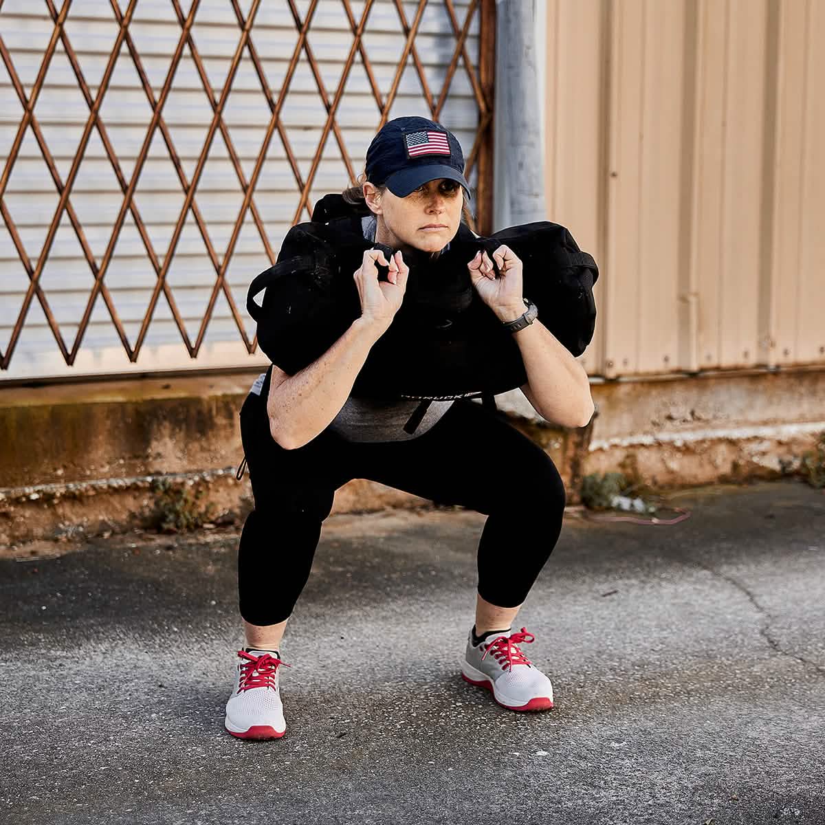 A person wearing a cap, black leggings, and sneakers performs a squat while lifting the GORUCK Ruck Clubs Starter Kit - Heavy on their shoulders. They're rucking outdoors, training near a closed metal gate as if preparing for an intense GORUCK event.