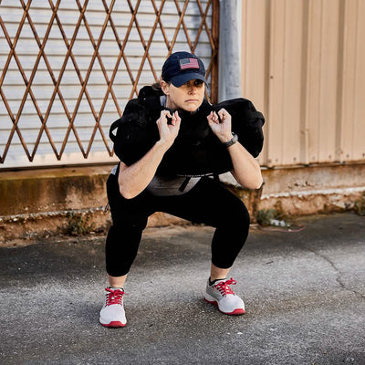 A person wearing a cap, black leggings, and sneakers performs a squat while lifting the GORUCK Ruck Clubs Starter Kit - Heavy on their shoulders. They're rucking outdoors, training near a closed metal gate as if preparing for an intense GORUCK event.