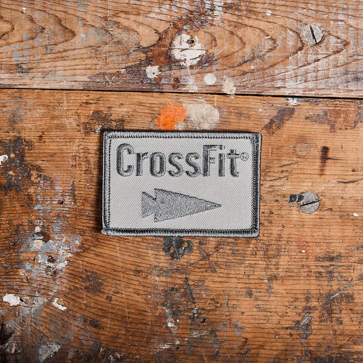 Resting on a rustic, paint-splattered wooden surface is the Patch - GORUCK x CrossFit, an embroidered piece by GORUCK-DEMO. This rectangular patch features the word CrossFit and an arrow symbol on a gray background with a black border, offering a rugged addition to any collection.