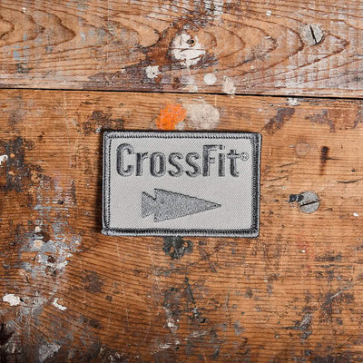 Resting on a rustic, paint-splattered wooden surface is the Patch - GORUCK x CrossFit, an embroidered piece by GORUCK-DEMO. This rectangular patch features the word CrossFit and an arrow symbol on a gray background with a black border, offering a rugged addition to any collection.
