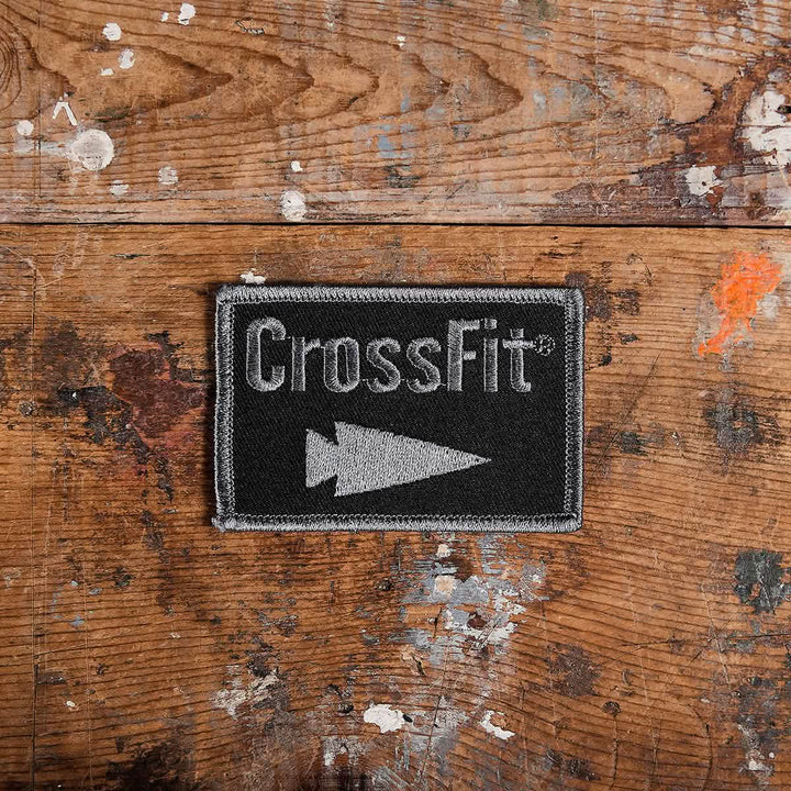 A GORUCK x CrossFit patch, crafted by GORUCK-DEMO and showcasing an arrow symbol in black and gray, is placed on a worn wooden surface adorned with colorful paint splatters.