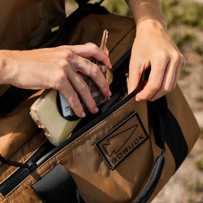 Close-up of hands placing a phone and keys into the brown Kit Bag - X-PAC, featuring a "GORUCK" patch and crafted from durable X-PAC VX21 material inspired by Special Forces gear.