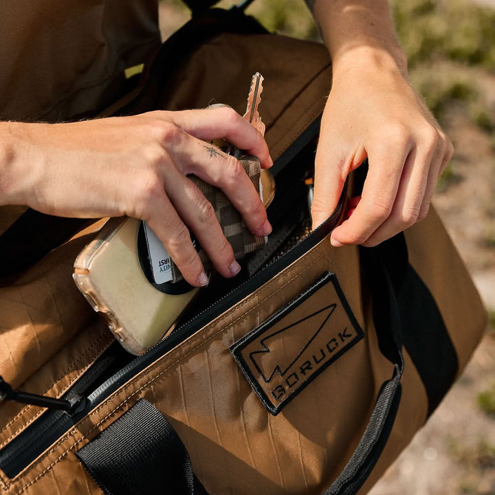 Close-up of hands placing a phone and keys into the brown Kit Bag - X-PAC, featuring a "GORUCK" patch and crafted from durable X-PAC VX21 material inspired by Special Forces gear.
