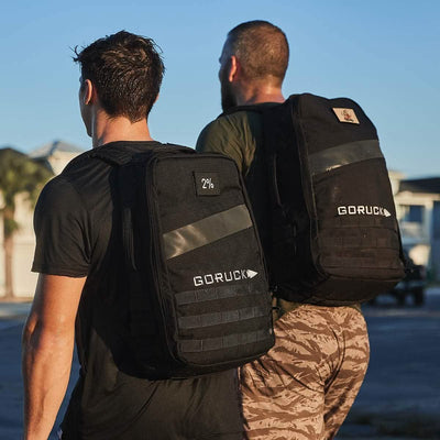 Two men walking outdoors, each sporting a black GORUCK Rucker 4.0 backpack. One wears a black shirt while the other dons a green shirt with camo pants. They stride through a sunlit, urban setting with buildings in the background, fully embracing the spirit of rucking.