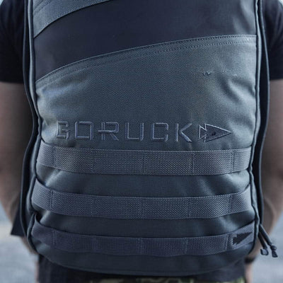 Close-up of a person wearing a grey and black GORUCK Rucker 4.0 backpack featuring MOLLE webbing. The GORUCK brand name is prominently displayed on the front, making it perfect for rucking enthusiasts.