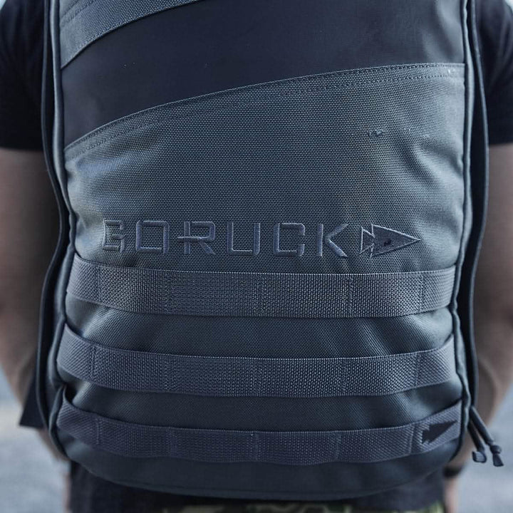 Close-up of a person wearing a grey and black GORUCK Rucker 4.0 backpack featuring MOLLE webbing. The GORUCK brand name is prominently displayed on the front, making it perfect for rucking enthusiasts.