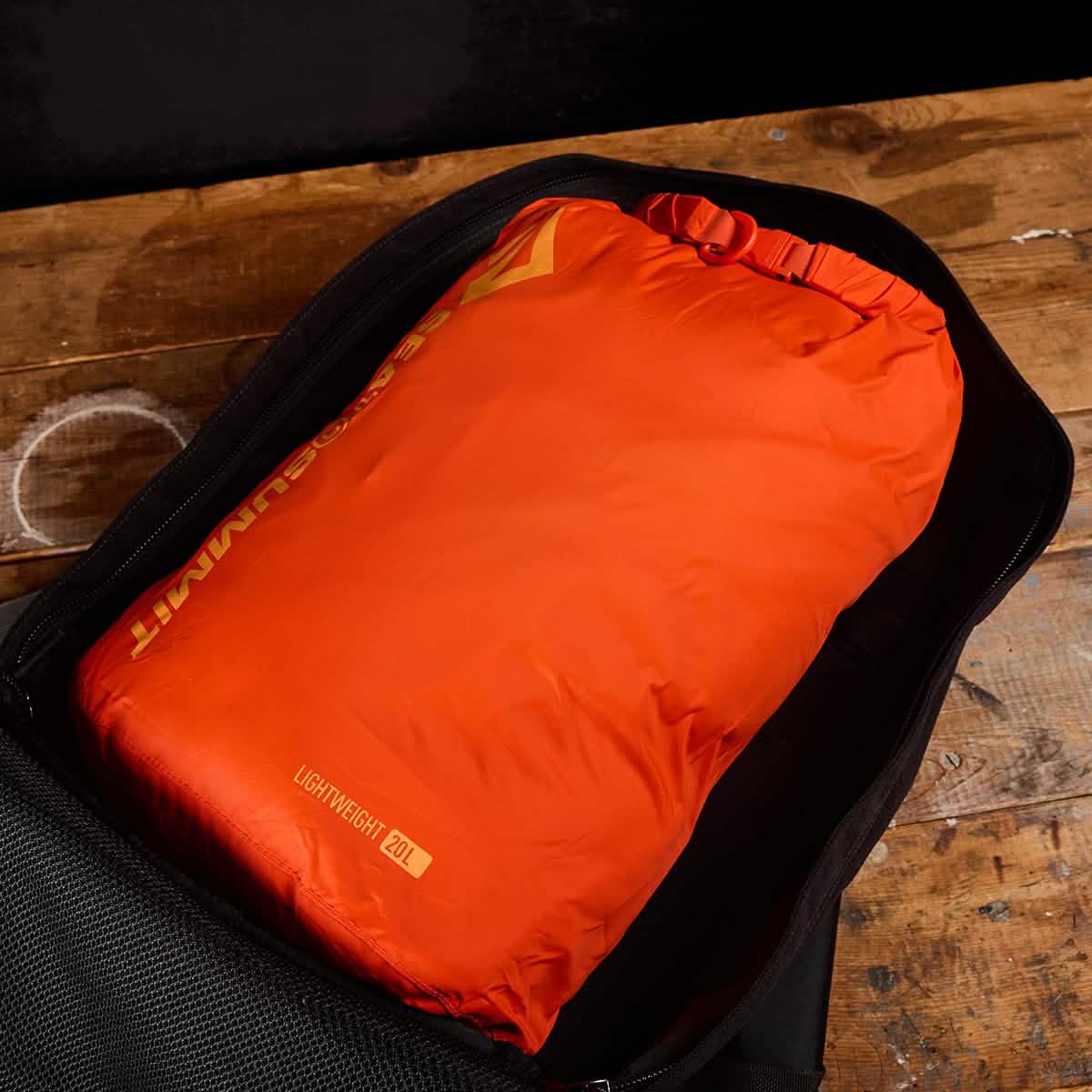 An orange Dry Sack from Sea to Summit, crafted with lightweight nylon, is neatly packed inside an open black backpack on top of a wooden surface. The sack is labeled with "Lightweight 62L" and includes a waterproof closure.