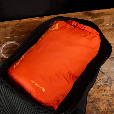 An orange Dry Sack from Sea to Summit, crafted with lightweight nylon, is neatly packed inside an open black backpack on top of a wooden surface. The sack is labeled with "Lightweight 62L" and includes a waterproof closure.
