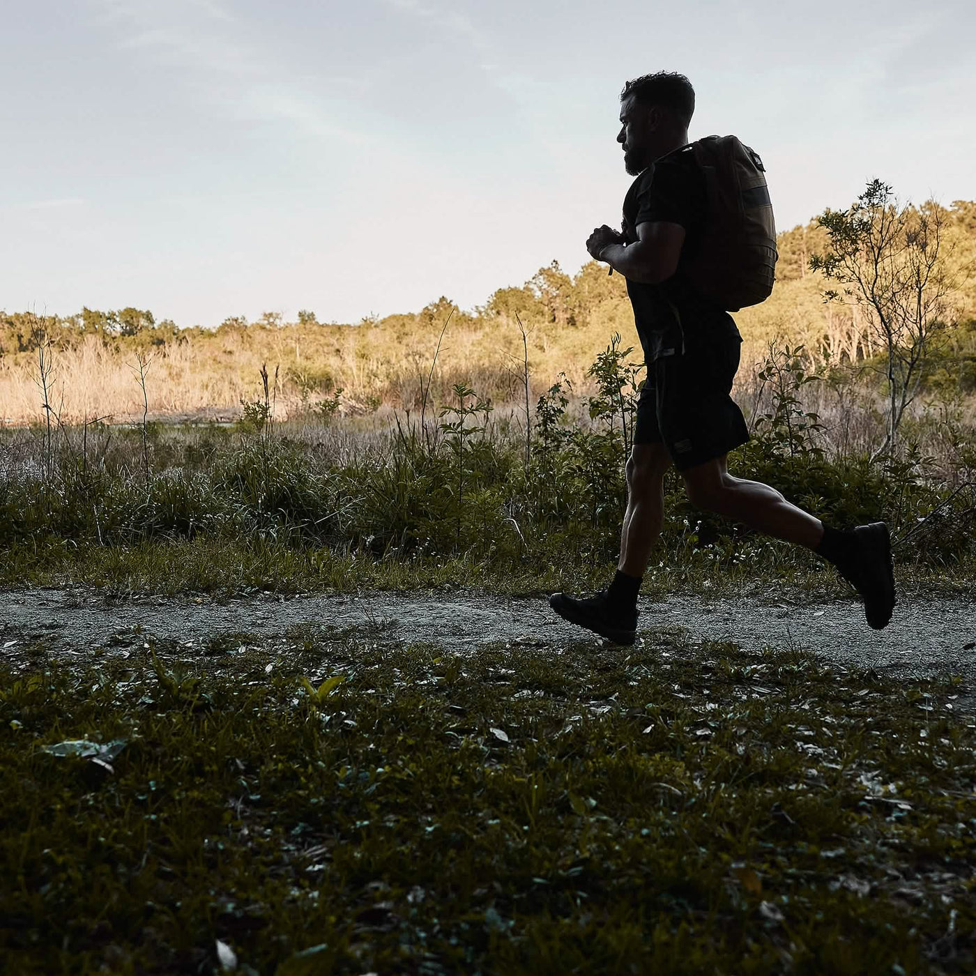 A man runs along a dirt path in a scenic outdoor setting, carrying the GORUCK Mackall backpack in Black + Forged Iron, resembling Special Forces training. Trees and bushes line the path under a clear sky, indicating early morning or late afternoon light.
