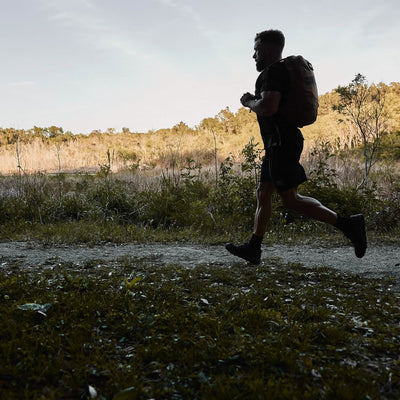 A man runs along a dirt path in a scenic outdoor setting, carrying the GORUCK Mackall backpack in Black + Forged Iron, resembling Special Forces training. Trees and bushes line the path under a clear sky, indicating early morning or late afternoon light.