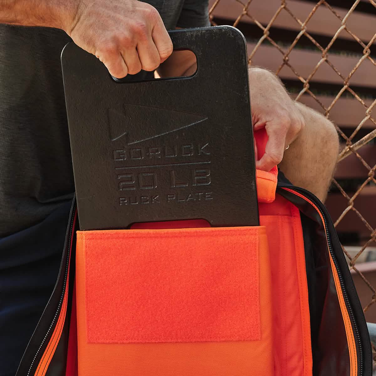An individual demonstrates their commitment to the demanding activity of rucking by placing a black GORUCK Ruck Clubs Starter Kit - Light into an orange backpack, set against the backdrop of a chain-link fence.