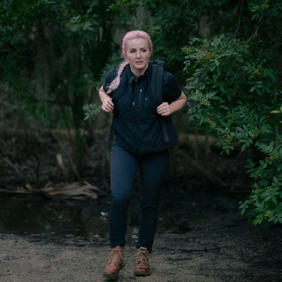 A person with pink hair, dressed in black and carrying a GR1 USA - X-PAC waterproof rucksack, walks through a wooded area.