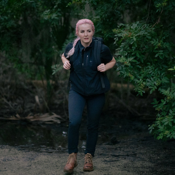 A person with pink hair, dressed in black and carrying a GR1 USA - X-PAC waterproof rucksack, walks through a wooded area.