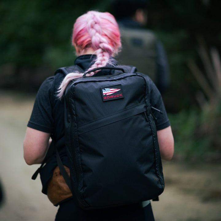 A person with pink braided hair strides along a dirt path, sporting a GR1 USA - X-PAC rucksack.